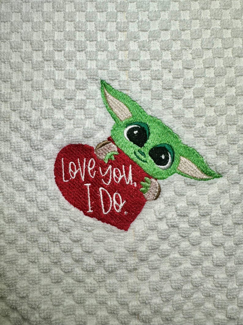 May include: A white kitchen towel with a green and red embroidered design. The design features a cartoon character holding a red heart with the text "Love you. I Do."