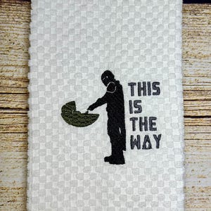 May include: White kitchen towel with an embroidered image of a Star Wars character holding a baby Yoda. The text "This is the way" is embroidered below the image.