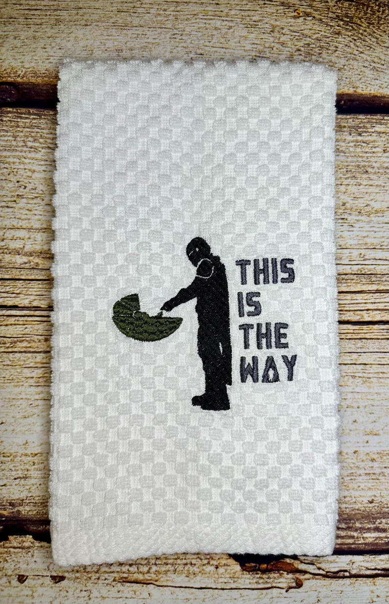 May include: White kitchen towel with an embroidered image of a Star Wars character holding a baby Yoda. The text "This is the way" is embroidered below the image.