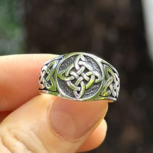May include: A silver-colored ring with a Celtic knot design. The ring features a central knot surrounded by intricate interwoven patterns. The ring is held up against a blurred green background, highlighting the detailed craftsmanship.