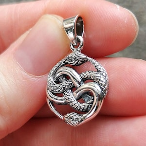 May include: A silver pendant featuring two intertwined snakes with detailed scales. The pendant has a loop at the top for attaching to a chain.