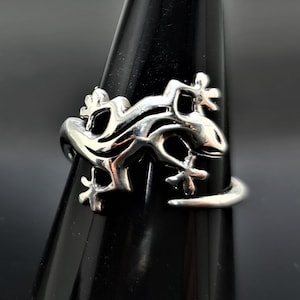 May include: A silver-colored ring featuring a lizard design. The ring is displayed on a black stand, showcasing the intricate details of the lizard's form. The lizard's body wraps around the finger, with its legs and tail extending.