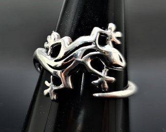 Puede incluir: Un anillo plateado con un diseño de lagarto. El anillo se exhibe sobre un soporte negro, mostrando los intrincados detalles de la forma del lagarto. El cuerpo del lagarto se envuelve alrededor del dedo, con sus patas y cola extendiéndose.
