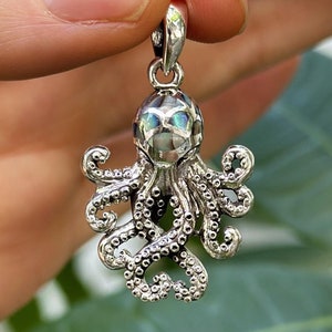 Peut inclure: Pendentif argenté en forme de pieuvre, aux détails complexes. La pieuvre a un corps et des tentacules texturés, avec une tête irisée. Le pendentif est suspendu à une petite boucle, prêt à être porté.