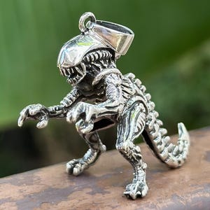Könnte beinhalten: Ein detaillierter Silberanhänger in Form einer Alien-Kreatur. Der Anhänger zeigt detaillierte Details des Kopfes, des Körpers und des Schwanzes der Kreatur. Der Alien steht mit ausgestreckten Armen. Der Anhänger ist als Halskette konzipiert.