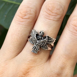 Könnte beinhalten: Ein silberner Ring mit einem detaillierten Engelsdesign mit ausgebreiteten Flügeln.