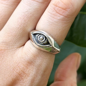Puede incluir: Un anillo de plata con un diseño de ojo. El anillo presenta un ojo detallado con un iris en espiral y una superficie texturizada. El anillo se lleva en un dedo, con un fondo verde.