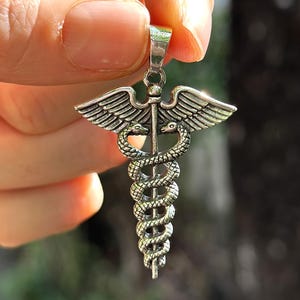 Puede incluir: Colgante plateado que presenta el símbolo del Caduceo, un bastón con dos serpientes enrolladas, coronado por alas. El colgante se sostiene sobre un fondo borroso, mostrando los intrincados detalles del diseño.
