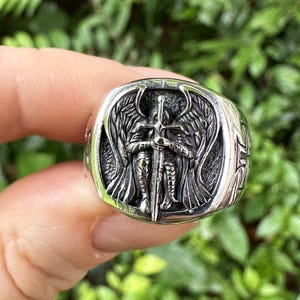 Könnte beinhalten: Ein silberner Ring mit einem detaillierten Design eines Engels, der ein Schwert hält. Der Engel hat Flügel und trägt eine Rüstung. Der Ring ist ein Statement-Piece und wäre ein tolles Geschenk für jemanden, der Engel liebt.