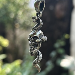 May include: Silver pendant necklace featuring a skull with a snake wrapped around it. The skull has detailed facial features, and the snake is coiled around the skull. The pendant hangs from a delicate silver chain, creating a gothic or biker-inspired aesthetic.