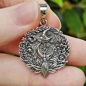 May include: Silver pendant with a witch hat, crescent moon, stars, and floral details. The pendant has a detailed, antique finish and is suspended from a simple bail. The design is circular, with a witch's broom at the bottom.