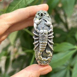 Puede incluir: Una pulsera de plata con un diseño de escorpión detallado. El escorpión está en relieve y tiene una superficie texturizada.