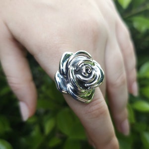 May include: A silver ring featuring a detailed rose design. The ring is worn on a finger and the rose is the focal point of the image.