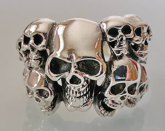 May include: A silver ring featuring a detailed design of multiple skulls. The ring has a polished finish, with the skulls arranged around the band. The skulls are intricately carved, showcasing the eye sockets, teeth, and facial features.
