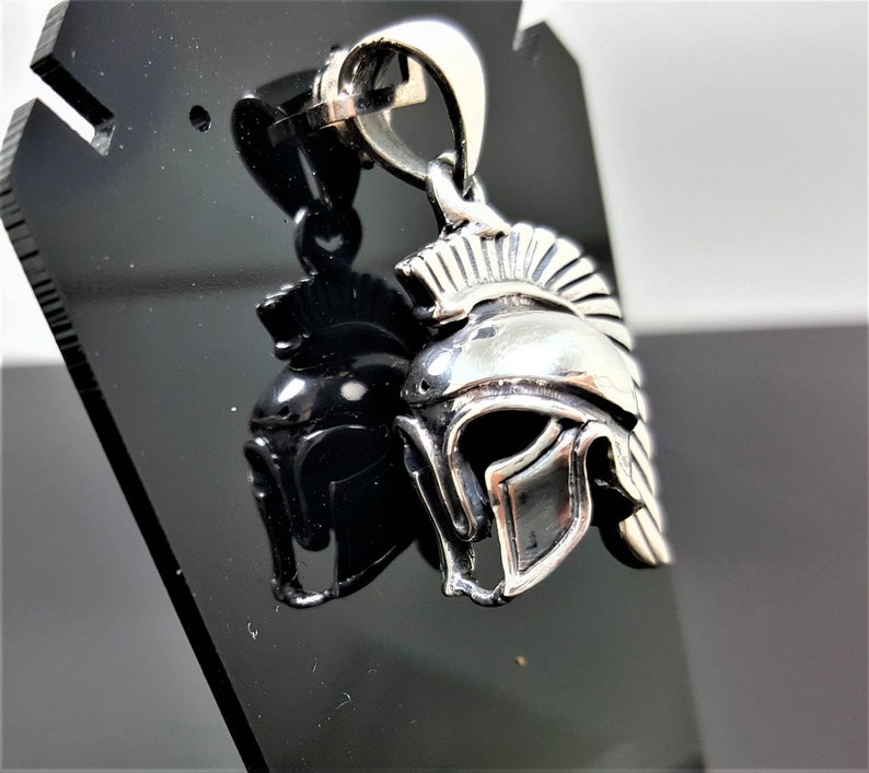 May include: Silver pendant in the shape of a Roman helmet with a plume on top. The pendant is on a black display stand.