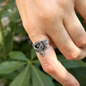 May include: A silver ring featuring a detailed angel design with outstretched wings. The angel is depicted with a delicate face and flowing hair.