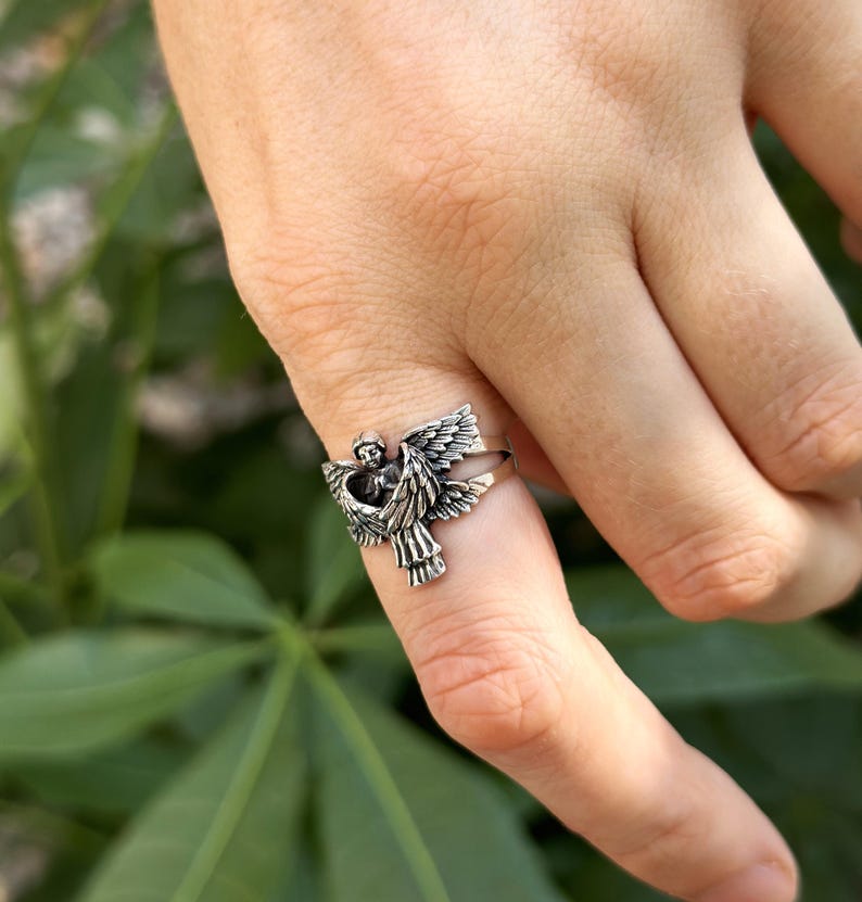 May include: A silver ring featuring a detailed angel design with outstretched wings. The angel is depicted with a delicate face and flowing hair.