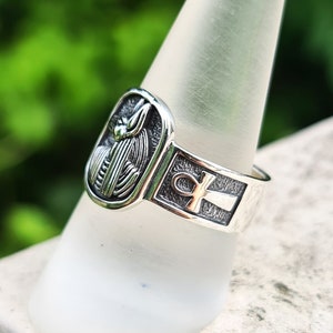 May include: Silver ring with an ankh symbol and a detailed depiction of an Egyptian god. The ring is on a white display stand.