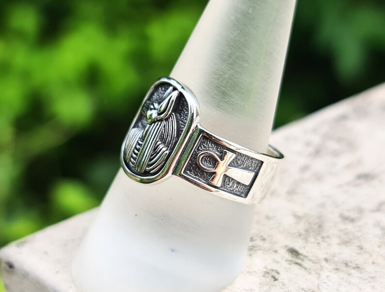 May include: Silver ring with an ankh symbol and a detailed depiction of an Egyptian god. The ring is on a white display stand.