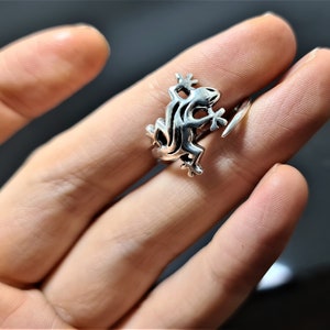 May include: A silver-colored ring featuring a stylized frog design. The frog is depicted in a detailed, three-dimensional form, with its legs and body clearly defined. The ring is worn on a finger, showcasing the intricate details of the frog.
