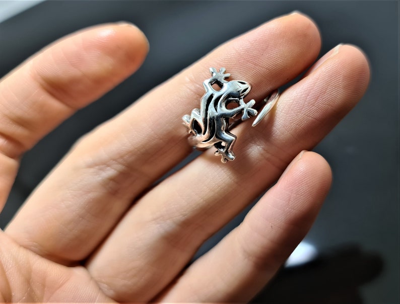 May include: A silver-colored ring featuring a stylized frog design. The frog is depicted in a detailed, three-dimensional form, with its legs and body clearly defined. The ring is worn on a finger, showcasing the intricate details of the frog.