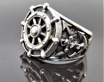 May include: A silver ring with a detailed design of a ship's wheel and anchor. The ring is made of metal and has a textured finish.