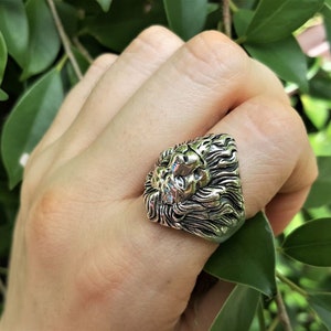 May include: A silver ring with a detailed lion head design. The ring is worn on a finger.