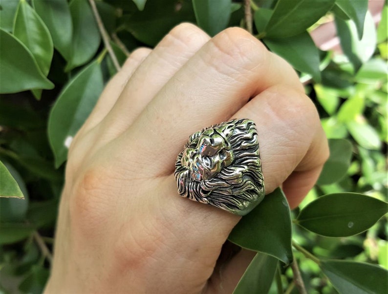 May include: A silver ring with a detailed lion head design. The ring is worn on a finger.