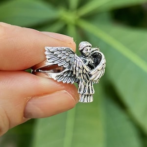 May include: A silver ring featuring a detailed sculpture of an angel with wings spread wide. The angel is depicted with a serene expression and flowing hair.