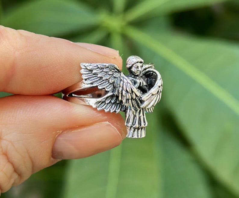 May include: A silver ring featuring a detailed sculpture of an angel with wings spread wide. The angel is depicted with a serene expression and flowing hair.