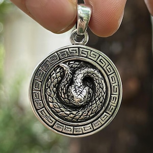 Puede incluir: Pendiente de plata con un diseño de serpiente detallado y un borde de llave griega. La serpiente está enroscada y su cabeza está mirando hacia adelante.