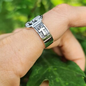 May include: A silver ring with an ankh symbol and a textured design. The ring is worn on a finger.