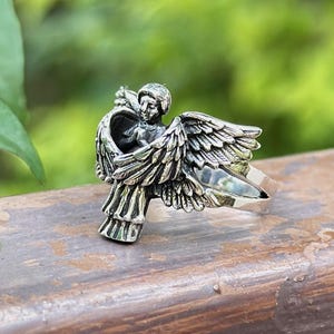 May include: A silver ring featuring a detailed sculpture of an angel with outstretched wings.