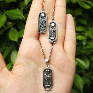 Puede incluir: Conjunto de collar y pendientes de plata con un colgante de escarabajo egipcio y pendientes a juego. El colgante y los pendientes presentan un escarabajo con alas, un disco solar y jeroglíficos.