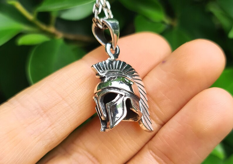 May include: Silver pendant necklace featuring a detailed depiction of a Spartan helmet. The helmet is a classic symbol of strength and courage.