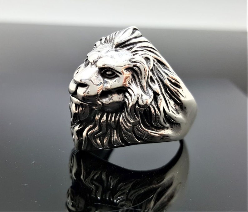 May include: A silver ring with a detailed lion head design. The lion's head is facing to the side and has a textured mane.