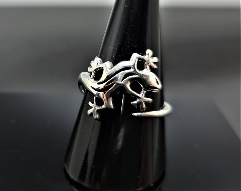 May include: A silver-colored ring featuring a lizard design. The ring is displayed on a black stand, showcasing the intricate details of the lizard's form. The lizard's body wraps around the finger, with its legs and tail extending.