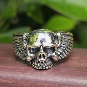 May include: A silver ring depicting a skull with detailed features and wings on either side. The ring's design is intricate, set against a blurred green and brown backdrop. The ring is a piece of jewellery.