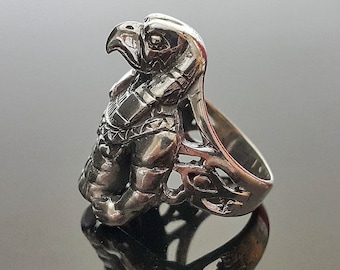 May include: A silver ring with a detailed design of a hawk or falcon head. The ring is made of metal and has a detailed design of the bird's head, with its beak open and its eyes looking forward. The ring is a statement piece and would be a great addition to any jewelry collection.