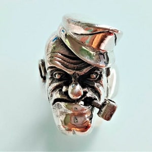 May include: A silver ring featuring a detailed sculpture of a man's face with a pipe in his mouth. The man is wearing a hat with a brim.