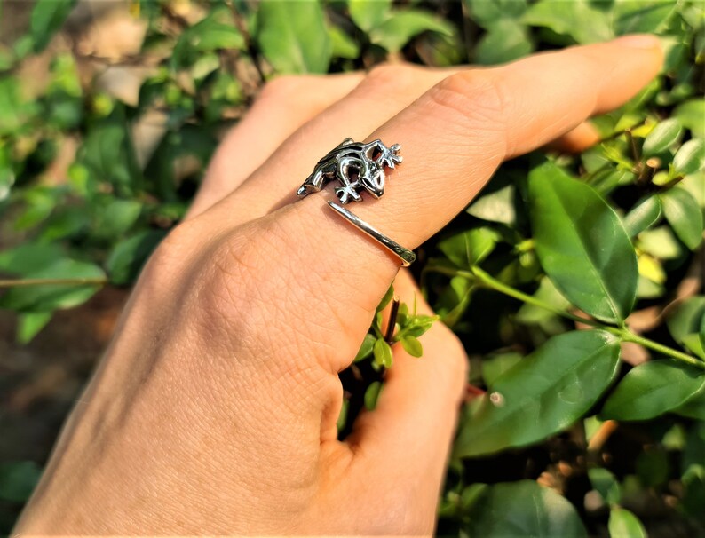 May include: A silver-toned frog ring. The ring features a detailed frog design wrapping around the finger. The frog is positioned on the top of the finger, with its legs and body visible. The ring is set against a backdrop of green foliage.