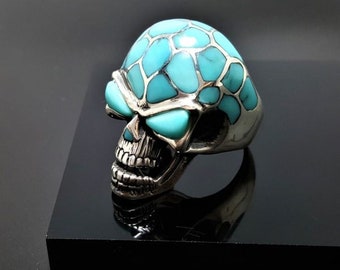 Può includere: Un anello in argento a forma di teschio con intarsi in turchese. Il teschio è dettagliato con un ghigno dentato e occhi turchesi.