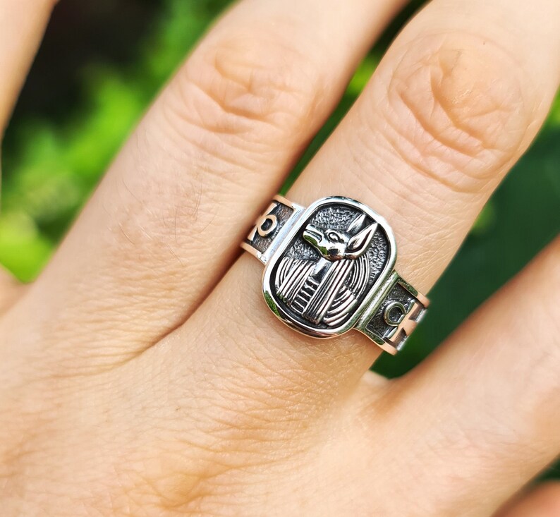 May include: Silver ring with an Anubis head design. The ring has a rectangular shape and is engraved with the Anubis head in the center. The ring is worn on a finger.