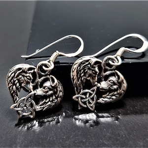 May include: Silver-toned earrings featuring a wolf design. Each earring showcases two wolves facing each other, with a Celtic knot detail. The earrings have a hook closure.