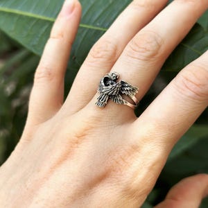 May include: A silver ring featuring a detailed angel design with outstretched wings. The angel is holding a heart in its hands.