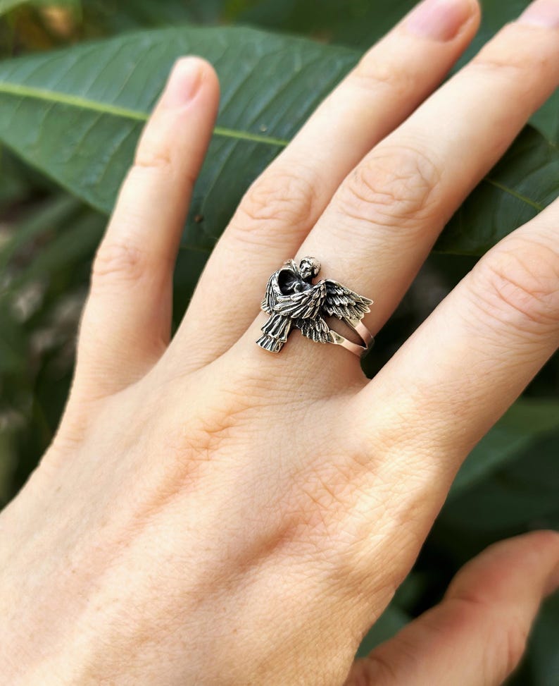 May include: A silver ring featuring a detailed angel design with outstretched wings. The angel is holding a heart in its hands.