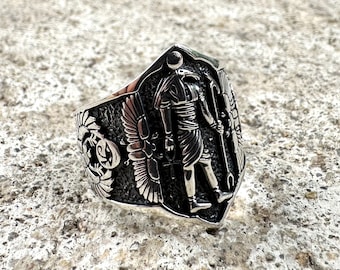 May include: A silver ring with an intricate design featuring an Egyptian god with wings and a staff. The ring is set against a textured background.