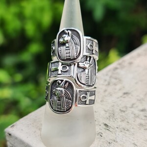 May include: Three silver rings stacked on a white display stand. Each ring features an Egyptian-inspired design with an ankh symbol and a depiction of an animal head.