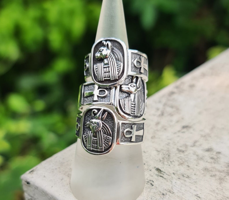 May include: Three silver rings stacked on a white display stand. Each ring features an Egyptian-inspired design with an ankh symbol and a depiction of an animal head.