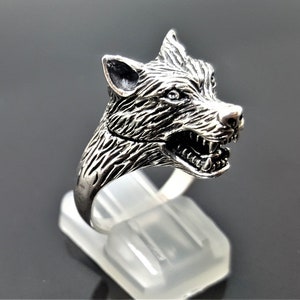May include: A silver ring featuring a detailed wolf head design. The wolf's mouth is open, revealing its teeth. The ring is displayed on a white stand.